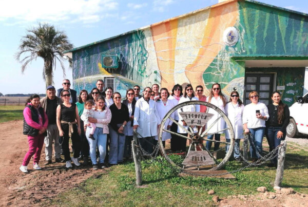 Grupo de personas, incluidos profesionales de la salud con túnicas blancas, posando frente a un edificio decorado con un colorido mural. En el centro de la imagen, se encuentra una estructura metálica con la inscripción 'Escuela N° 29 - Bellón'. Al fondo, se aprecia una palmera y un paisaje rural.