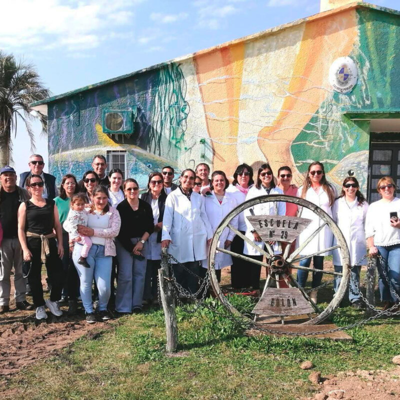 Grupo de personas, incluidos profesionales de la salud con túnicas blancas, posando frente a un edificio decorado con un colorido mural. En el centro de la imagen, se encuentra una estructura metálica con la inscripción 'Escuela N° 29 - Bellón'. Al fondo, se aprecia una palmera y un paisaje rural.